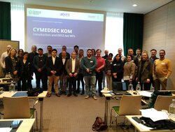 group of people standing in front of a projection slide that says "CYMEDSEC KOM. Introduction and EKFZ-led WPs". In the foreground one can see desks and chairs, laptops, paper, bags from the time the participants were sitting listening to the presentation.