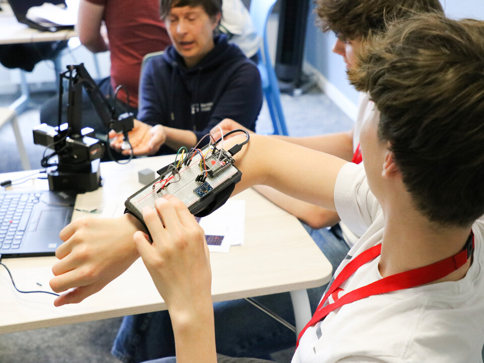 A person wearing a wrist-mounted circuit board with wires explores a project, while another individual interacts with a robotic arm. The room looks like is a classroom, conveying innovation and teamwork.