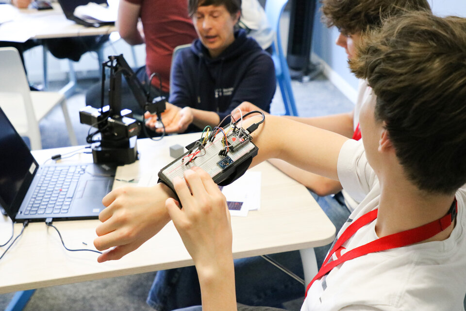 A person wearing a wrist-mounted circuit board with wires explores a project, while another individual interacts with a robotic arm. The room looks like is a classroom, conveying innovation and teamwork.