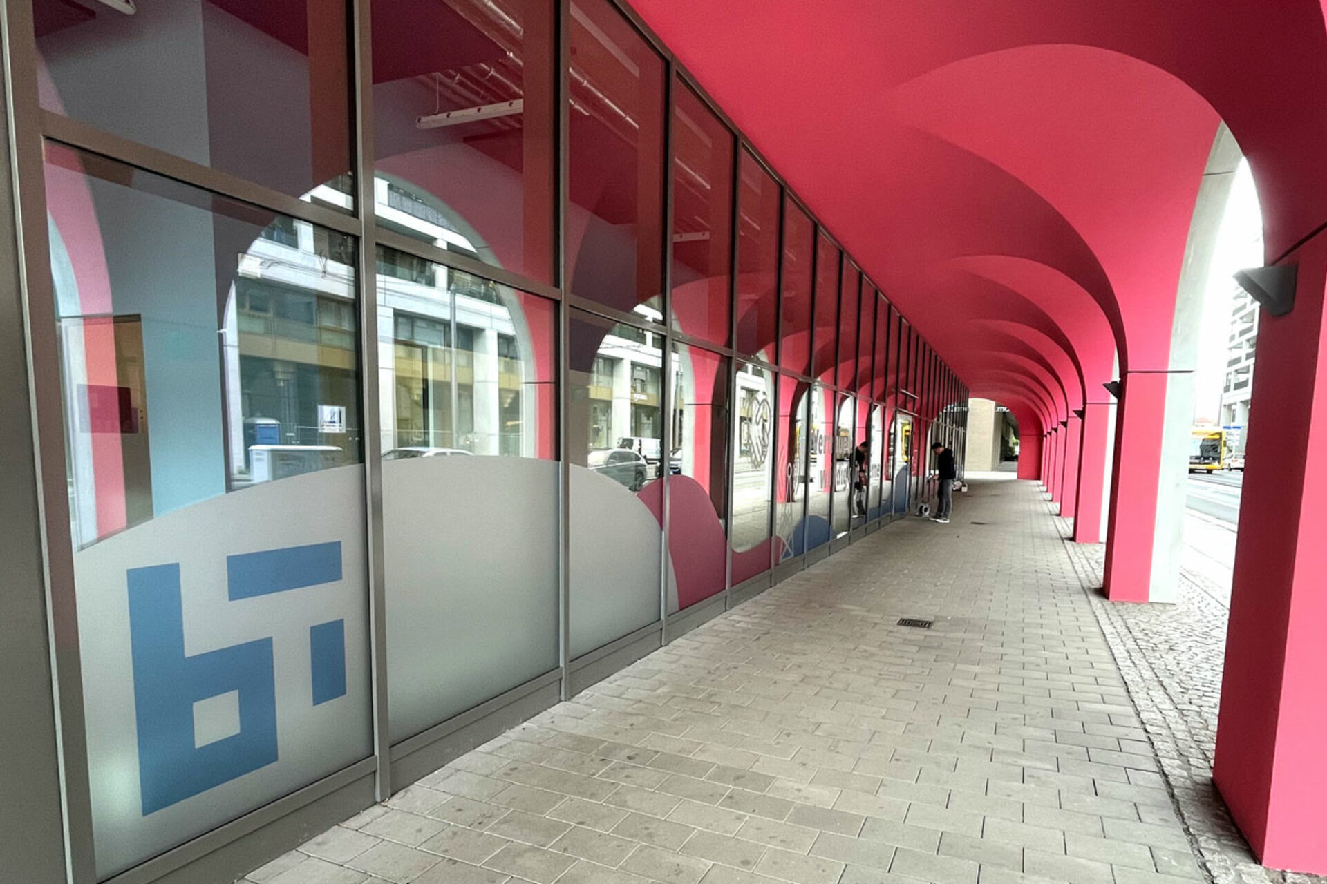 Langer überdachter Gehweg mit pinken Rundbögen und großen Fenstern mit halbtransparenten Folien und blauen grafischen Mustern. Links unten das Logo des Barkhausen Instituts.