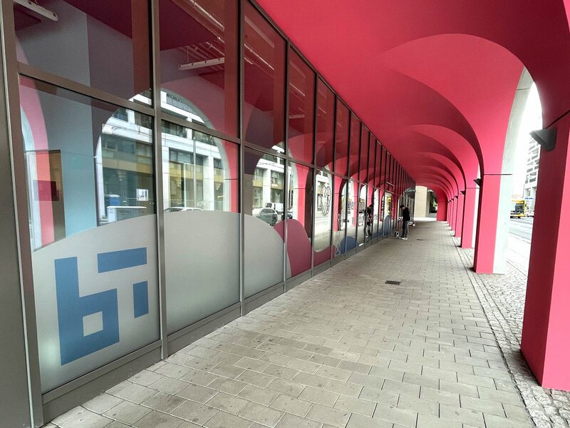 Langer überdachter Gehweg mit pinken Rundbögen und großen Fenstern mit halbtransparenten Folien und blauen grafischen Mustern. Links unten das Logo des Barkhausen Instituts.