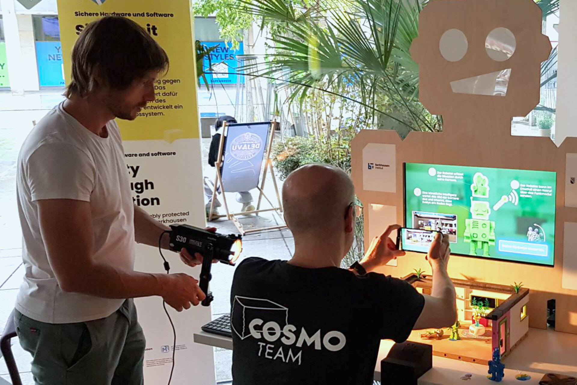 Ein Mann in einem schwarzen T-Shirt mit der Aufschrift „COSMO TEAM” filmt mit einem Smartphone. Ein weiterer Mann hält eine Lampe.