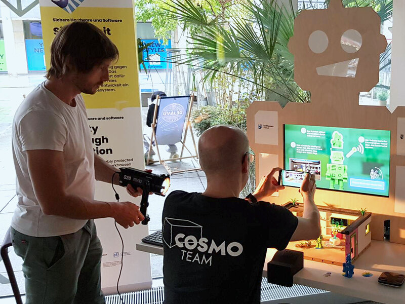 One man wearing a black T-Shirt saying "COSMO TEAM" is filming with a smartphone. One other man is holding a light.