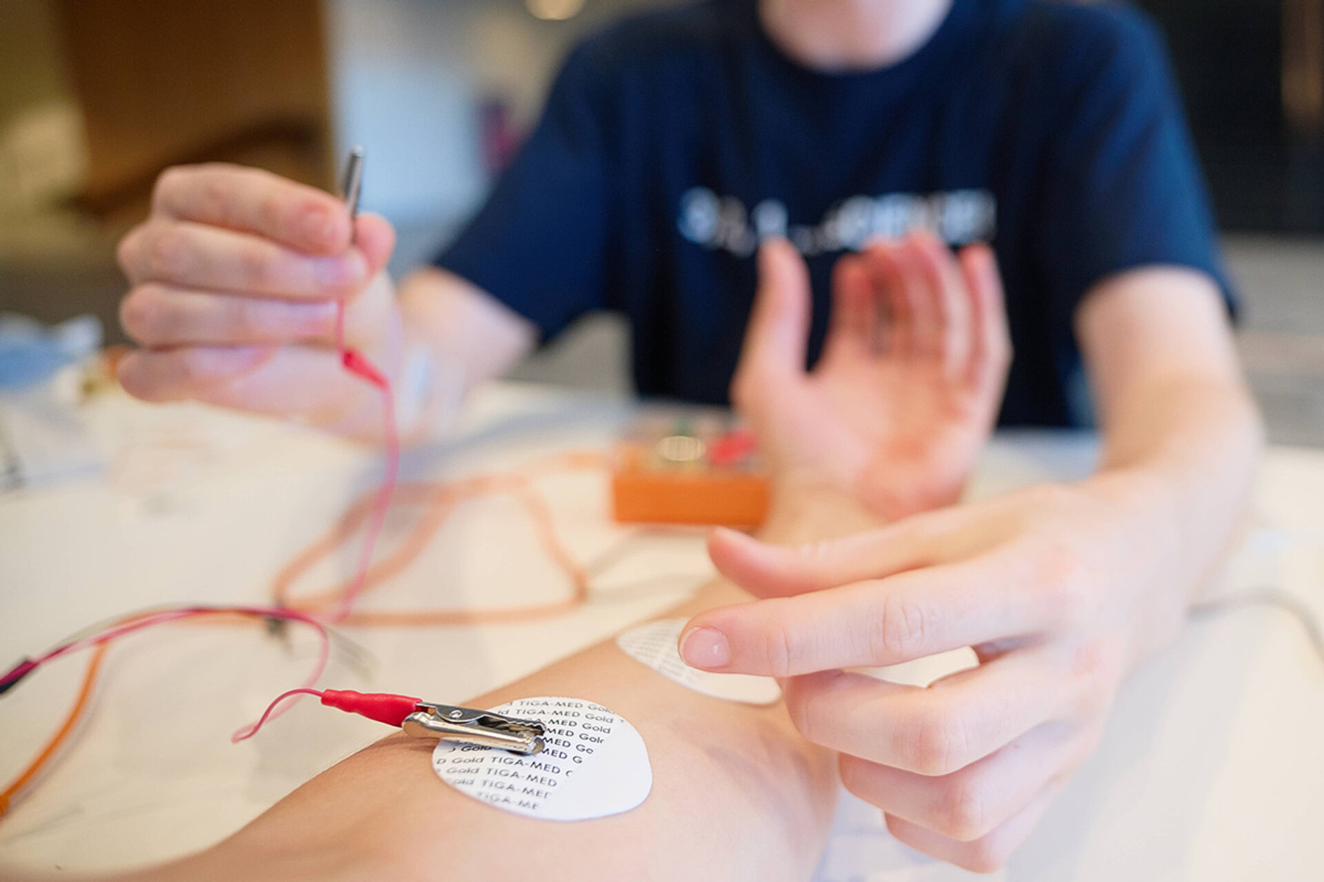 Nahaufnahme einer Person, die mit einer Elektrode auf dem Unterarm verbunden ist und deren Hand von einer anderen Person mit einer Klemme berührt wird