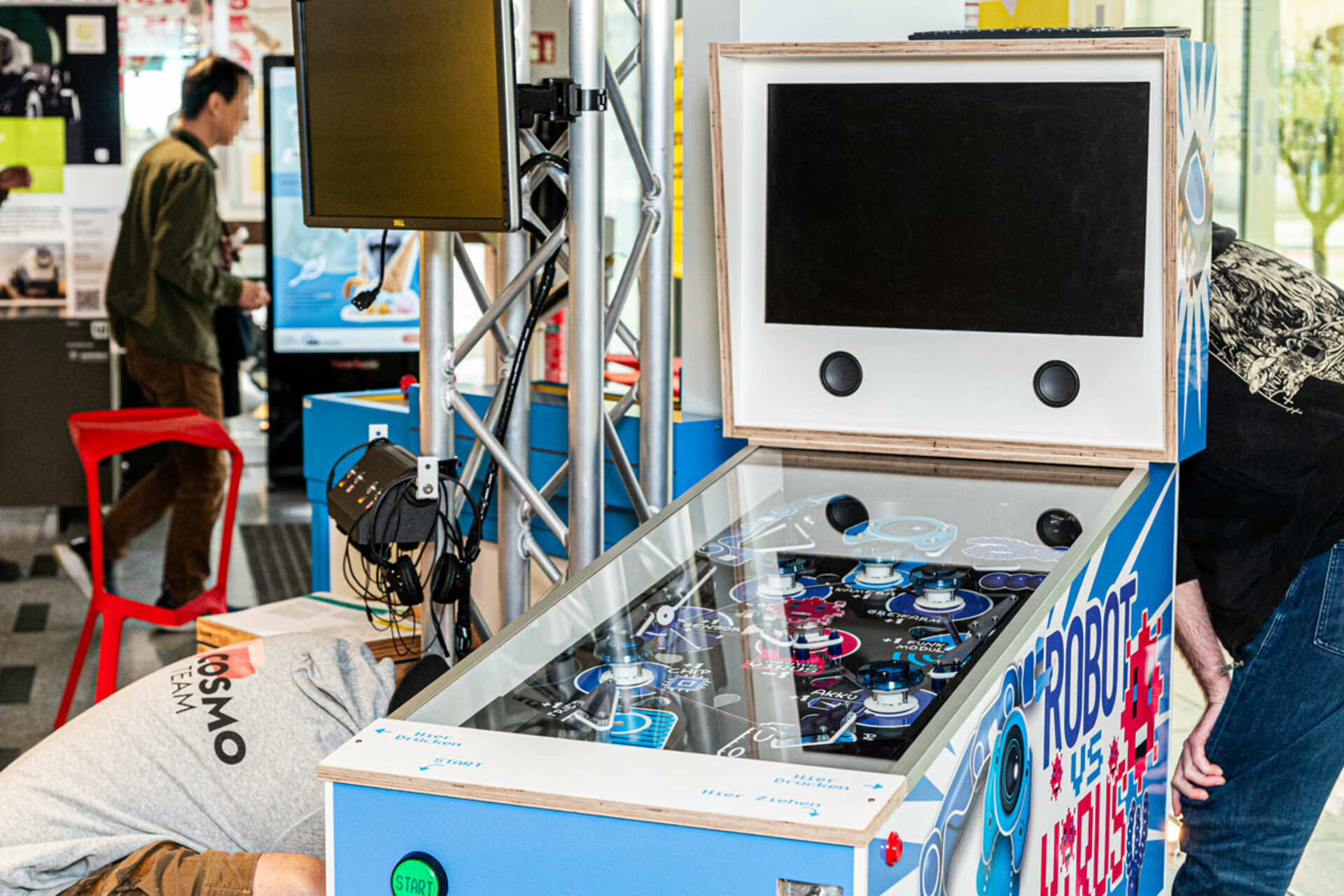 Interaktives Ausstellungsstück mit Joysticks und Knöpfen, beschriftet mit 'ROBOT VS. VIRUS', im Aufbau der Ausstellung 'Mensch, Roboter!' im COSMO Wissenschaftsforum.