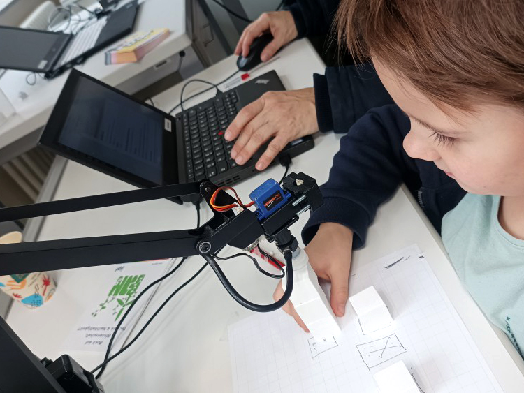 A child and a man sitting at a table, the man is working on a laptop and the child is interacting with a robot arm