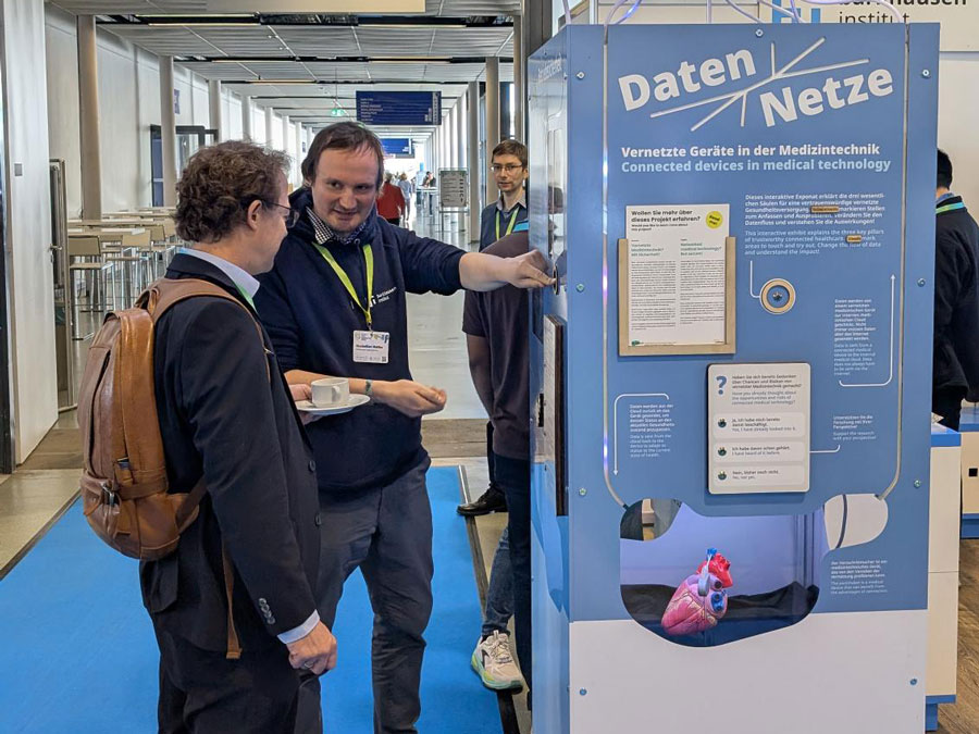 Zwei Männer im Gespräch vor einem blauen Informationsstand mit dem Titel 'Daten Netze' über vernetzte Geräte in der Medizintechnik, darunter ein Modell eines Herzens.