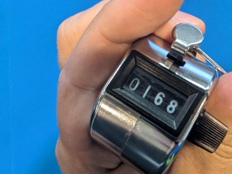 Hand hält einen silbernen mechanischen Zähler mit der Anzeige 0168 vor blauem Hintergrund.