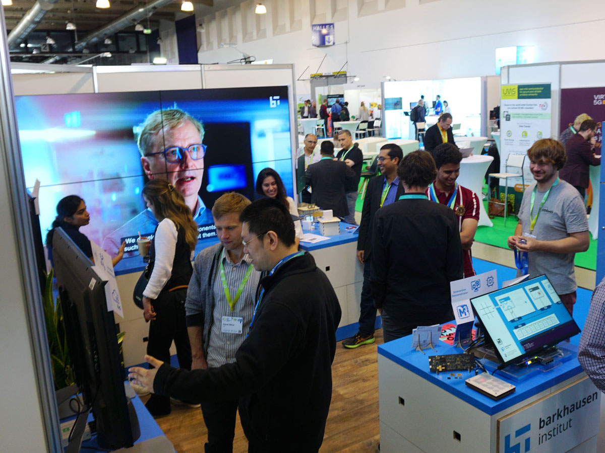 Menschen am Messestand des BI mit Bildschirmen und Informationsmaterial auf dem 6G Summit in der Messehalle Dresden
