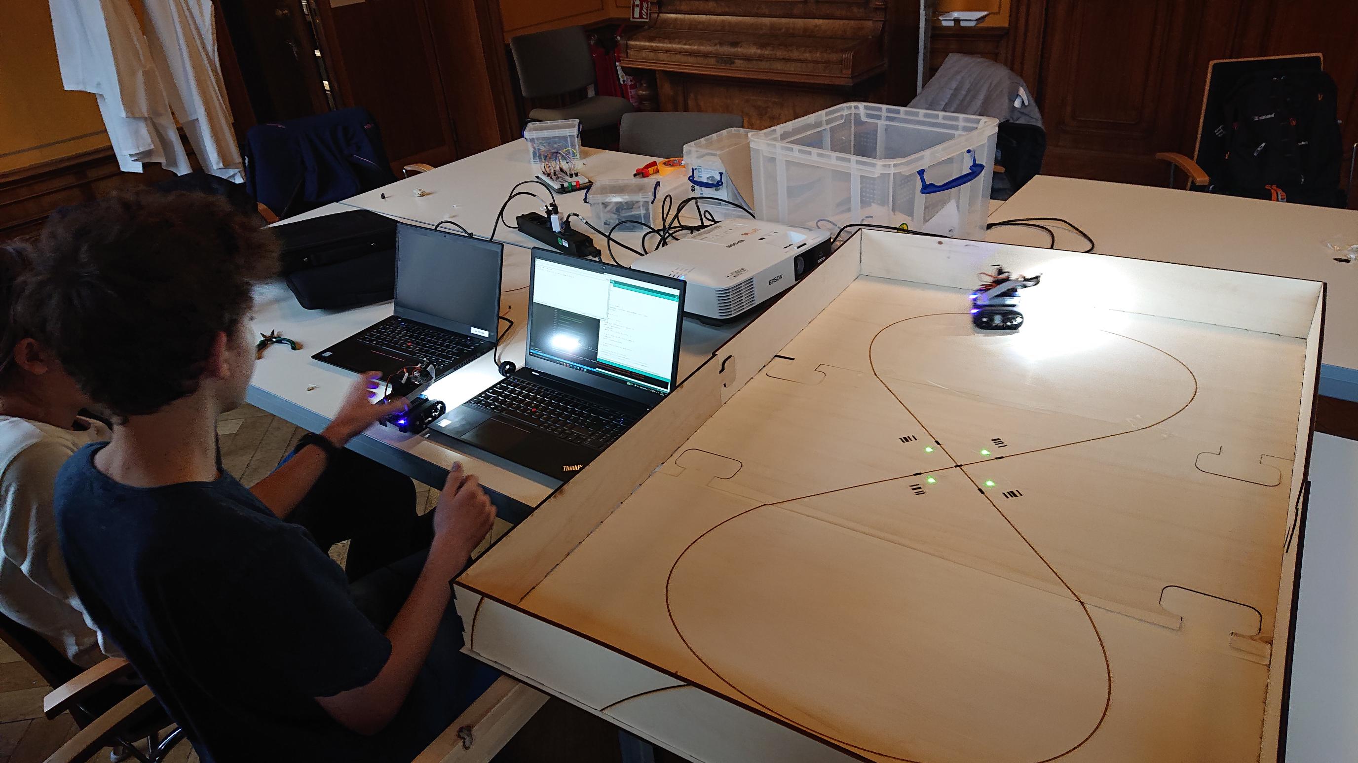 2 children sit at a table at a laptop. on the laptop screen one can see code. one kid is holding a pixybot, he just programmed. Another pixibot is on a cardboard racetrack right beside them.