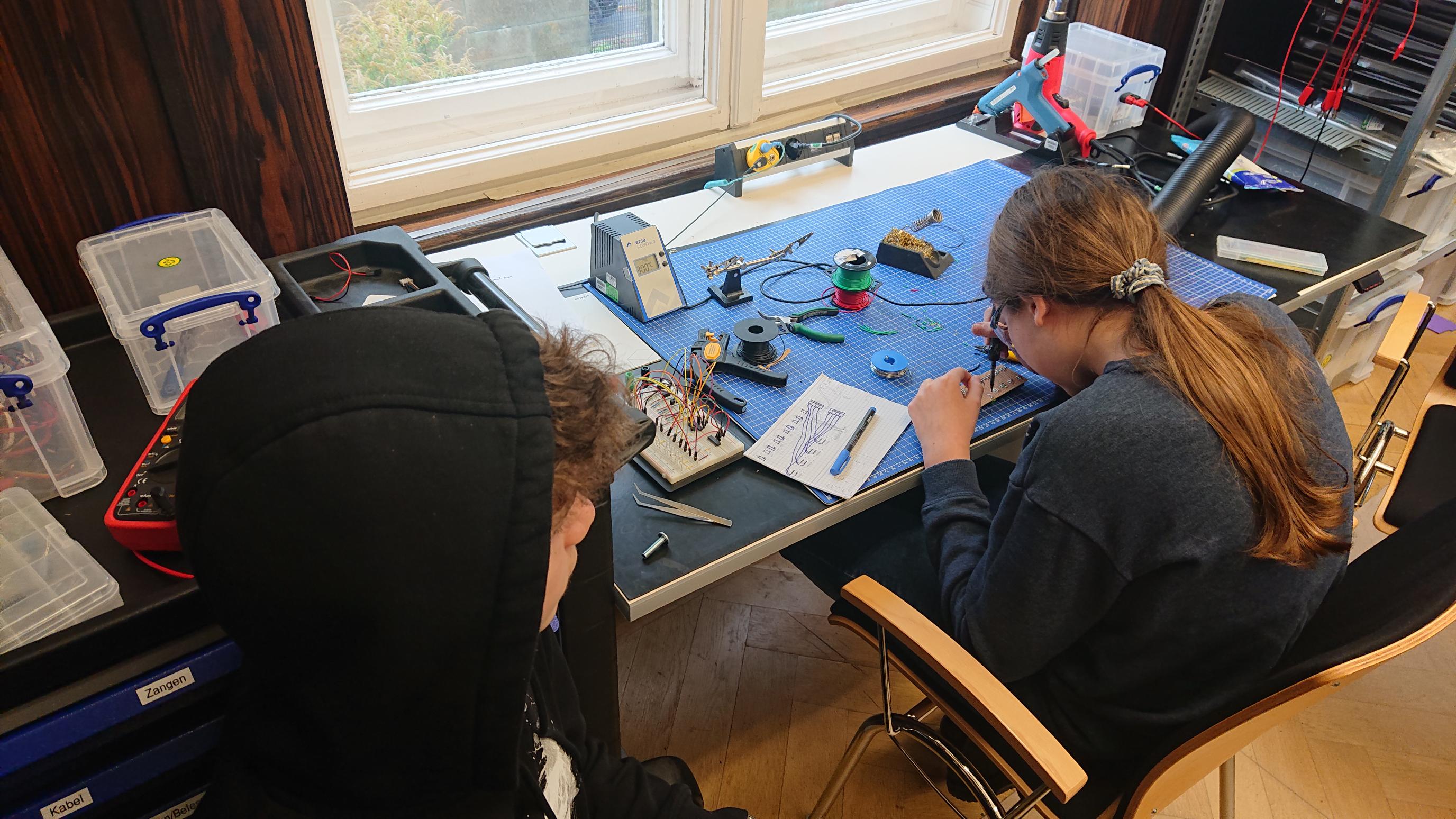 2 children sit at a table. one kid is soldering. the other one is watching.